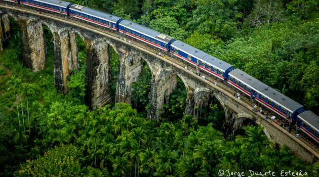 A beleza exuberante da floresta tropical de Ella, Sri Lanka, emoldura o Ponte dos Nove Arcos, enquanto um comboio azul e vermelho atravessa.