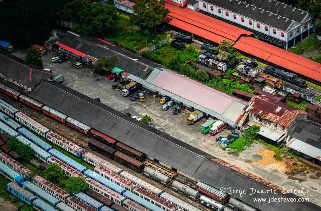 Vista aérea de um movimentado terminal ferroviário em Colombo, Sri Lanka, onde trens coloridos e antigos cruzam-se com edifícios de telhados vermelhos e cinzentos.