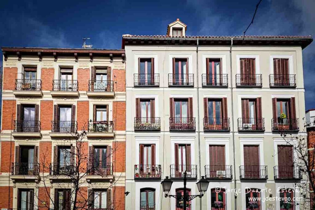 Fachada de edifícios residenciais tradicionais no bairro de La Latina, em Madrid, com varandas de ferro e persianas de madeira sob um céu azul. A arquitetura típica castiça para descobrir ao visitar Madrid.