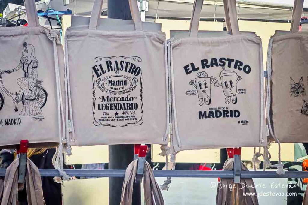 Sacos de pano ilustrados (tote bags) no Mercado El Rastro, Madrid; souvenirs típicos do bairro de La Latina.
