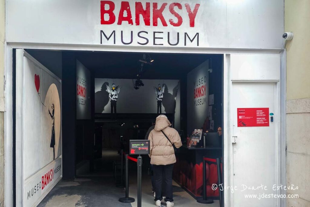 Fachada e entrada do Museu Banksy em Madrid, com a icónica obra da menina com o balão vermelho e visitantes na bilheteira.