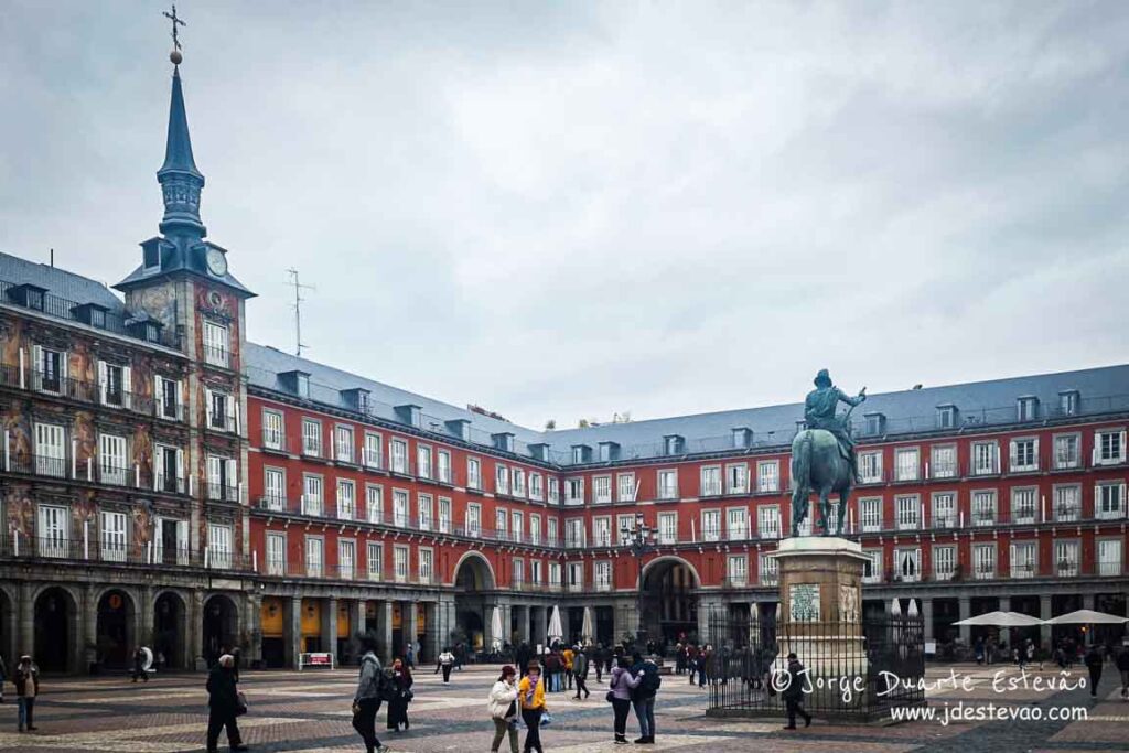Turistas caminham pela ampla Plaza Mayor de Madrid num dia nublado; pormenor da arquitetura de estilo madrileno com as suas torres e arcadas. Local obrigatório num roteiro por Madrid, Espanha.