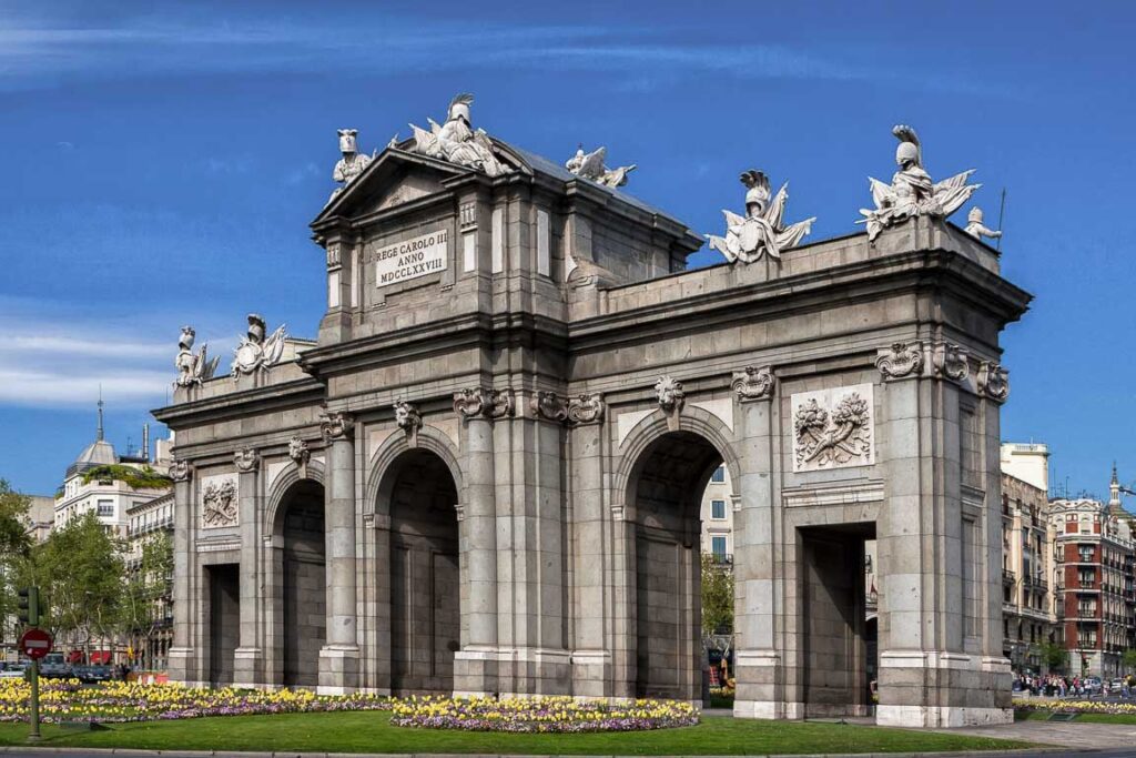 ista detalhada da Puerta de Alcalá, antiga porta de entrada da cidade de Madrid, destacando os seus arcos e esculturas em pedra. Arquitetura histórica no centro da capital espanhola.