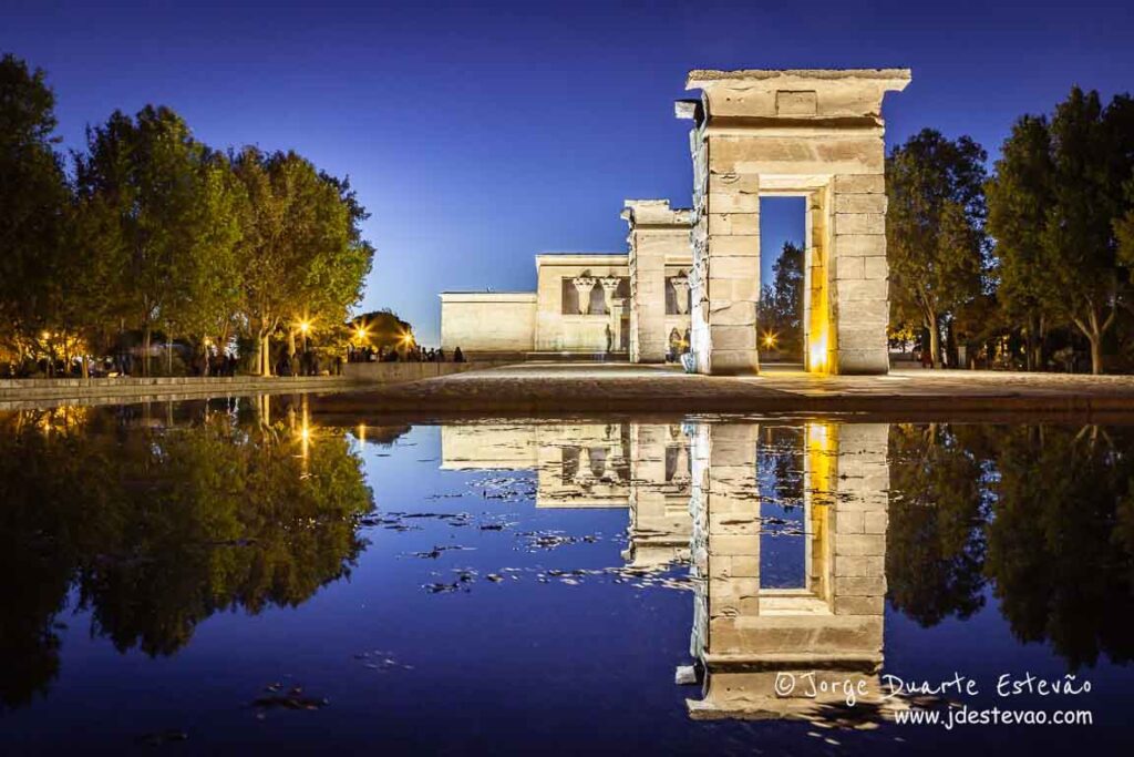 O milenar Templo de Debod iluminado ao entardecer com reflexo perfeito na água, um dos locais mais românticos e gratuitos para visitar em Madrid.